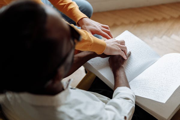 A mentor helps a visually impaired man read a Braille book indoors, fostering learning and support.