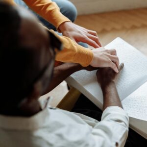 A mentor helps a visually impaired man read a Braille book indoors, fostering learning and support.